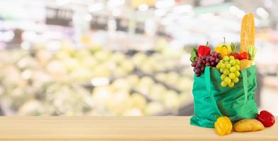 Fresh fruits and vegetables in reusable green shopping bag on wood table top with supermarket grocery store blurred defocused background with bokeh light photo