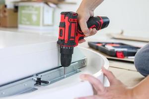 man assembling white table furniture at home using cordless screwdriver photo