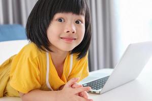 una pequeña estudiante asiática estudia en línea usando una computadora portátil en casa foto