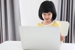 una pequeña estudiante asiática estudia en línea usando una computadora portátil en casa foto