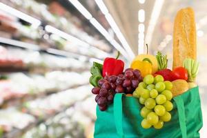 Fresh fruits and vegetables in reusable green shopping bag with supermarket grocery store blurred defocused background with bokeh light photo