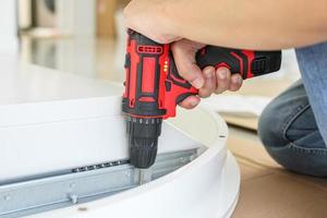 man assembling white table furniture at home using cordless screwdriver photo