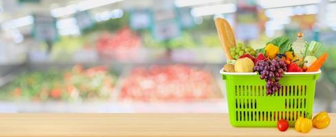 Shopping basket filled with fruits and vegetables on wood table with supermarket grocery store blurred defocused background with bokeh light photo