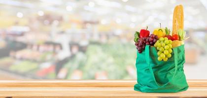 Fresh fruits and vegetables in reusable green shopping bag on wood table top with supermarket grocery store blurred defocused background with bokeh light photo