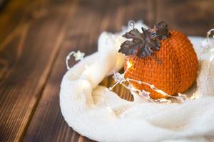 Orange pumpkin wrapped in a warm white plaid with lights of garlands on a wooden table with copyspace. Autumn mood, halloween, harvest, thanksgiving, home decor and cozy. Selective focus photo