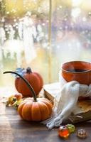 An orange mug in a scarf with hot tea, pumpkins, yellow dry maple leaves, a book on the windowsill, raindrops on the window - autumn mood photo