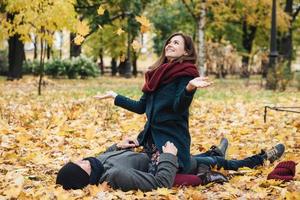 Beautiful female has fun with boyfriend, throws yellow leaves, sits on man, spend time together in autumn park, have happy expressions. Emotional girlfriend rejoice togetherness. Relationship concept photo