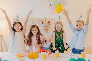Horizontal shot of happy positive children catch confetti, celebrate birthday together, raise arms, have good mood, play together, stand near festive table with present box, cake, party hats photo