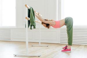 Horizontal shot of motivated woman does push up on ballet bare, warms up before aerobics, wears sportswear, stands in dance hall. Skillful pilates female instructor prepares muscles for training photo