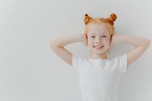 Indoor shot of glad smiling girl covers ears with hands, ignores noise, has two ginger buns, freckles, foolishes around, wears casual t shirt, isolated over white background, listens loud music photo