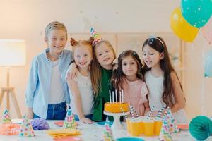 Horizontal shot of group of little children gather together to celebrate birthday, embrace and pose at camera, prepare for special occasion, stand near table with cake, paper cups, party caps photo