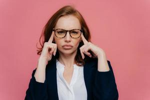 Portrait of intense beautiful lady touches temples with both hands, tries to recollect something important in mind, has serious concentrated look at camera, wears spectacles and elegant suit photo