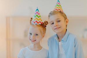 Horizontal shot of happy girl and boy wears cone party hats, celebrate birthday together, have good mood, wait for guests, pose indoor against blurred background. Children, holiday, celebration photo