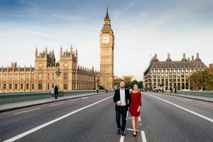 Female wears red elegant dress and man hold hands, have walk on Westmisnter bridge, admire Londons sights, have excursion, like travelling together. Young enthusiastic tourists in Great Britain photo