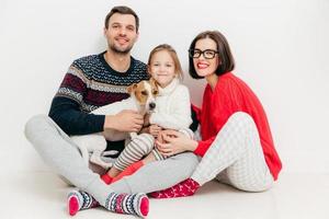Isolated shot of good looking female and male sit together with their daughter and jack russell terrier dog, embrace each other, isolated on white background. Family and togetherness concept photo