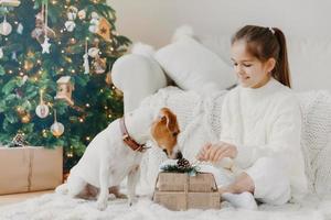 Horizontal shot of adorable small girl with pony tail, dressed in white winter sweater, sits crossed legs on floor, unpacks Christmas gift, beautiful decorated New Year tree near. Holiday traditions photo