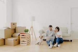 Affectionate married couple move in new home, relax on floor after unpacking things use laptop computer for searching home decoration ideas in internet their pet sits near, pose in purchased apartment photo