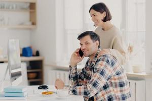 Indoor shot of pleased woman and man busy with discussing domestic expenses, cheerful husband in checkered shirt makes telephone call, caring wife touches shoulders, focuse in monitor of computer photo