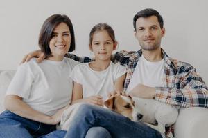 Portrait of pleasant looking mother, dad and small child, sleeping pet, pose all together at sofa against white wall, dressed in casual clothes, spend weekend at home, have friendly relationship photo