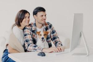 una pareja feliz y amistosa se sienta junto con un perro, reserva un hotel para un viaje futuro, usa el teclado y mira la computadora, chatea en línea, posa en un escritorio blanco, está conectado a wifi gratis, navega por Internet juntos