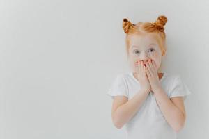 Horizontal shot of surprised red head girl covers mouth, watches exciting cartoon, has unexpected reaction, wears casual white t shirt, stands indoor, expresses wonder. Children and emotions concept photo