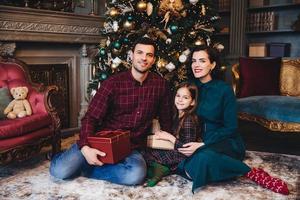 la foto interior de una familia joven tiene una buena relación, se sientan juntos en el piso cerca del árbol de año nuevo decorado, sostienen un regalo, se felicitan mutuamente por el próximo año nuevo y navidad. concepto de vacaciones