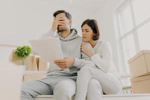 Dejected husband and wife manage finances, receive bill, face financial problem, have gloomy expressions, sit together in empty room, big window behind, cardboard boxes with personal stuff near photo