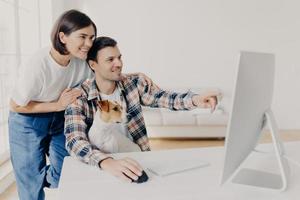 Happy man and his girlfriend consult about future project, point into monitor, pose at workplace together with dog in home interior, watch online tutorial video, spend time in modern apartment photo