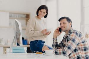 Photo of cheerful busy male freelancer uses mobile phone, discusses latest news, sits at desktop with papers and modern computer. Positive woman dressed in sweater and jeans, holds mug of drink