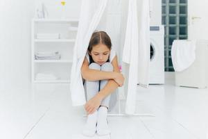 Lonely thoughtful sad small girl sits on floor in washing room near clothes dryer, wears trousers and white socks, washing machine, laundry basket and console in background, waits for parents photo