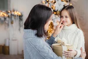 Beautiful female child with long hair, wears white warm sweater, looks in mothers eyes, glad to recieve present on Christmas, celebrate winter holidays in family circle. Happy mother and daughter photo