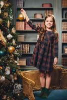 Pretty girl with charming smile stands on armchair, decorates New Year tree alone, has good mood, feels proud to do it by herself. Small kid prepares for Christmas. Childhood and holidays concept photo