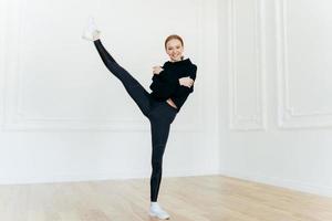 Fit satisfied red haired female raises leg, crosses hands over chest, demonstrates nice flexibility and resilience, wears black hoody and leggings, stands in fitness studio, has gymnastics exercises photo