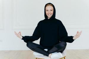 Horizontal shot of optimistic woman practices yoga, meditates on chair, sits in lotus pose, wears black sweatshirt, white sportshoes, has glad expression, tries to relax after hard working day photo