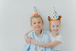 Lovey red haird girl embraces with love her older brother, congratulates with birthday, wear cone party hats, have good mood on party, isolated over white background, have friendly relationship photo