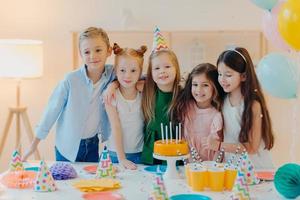 Friendly children embrace while pose near festive table, blow candles on cake, have party mood, celebrate birthday or special occasion, have joyful expressions. Childhood, fun and entertainment photo