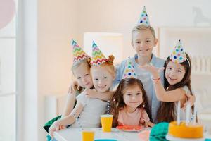 Indoor shot of happy kids celebrate party with falling confetti, wear cone party hats, pose near festive table with cakes, embrace and pose together. Childrens birthday photo