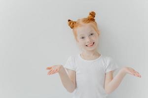 Pretty small female kid with double foxy buns spreads palms sideways, looks happily at camera, has green eyes, freckled skin, appealing look, wears casual white t shirt, poses indoor. Childhood photo