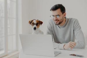 Busy man works freelance, watches training video online on laptop computer wears transparent glasses works together with jack russell terrier pose in home office read news online. Businessman with dog photo