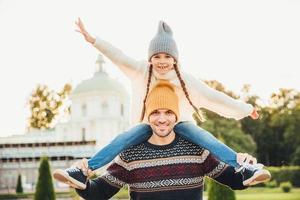 Horizontal portrait of affectionate father gives piggy back ride to his daughter outside, play together, have fun. Happy little charming girl spends weekends with father, waves excitedly with hand photo