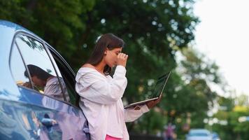 la mujer se apoya en el auto mientras sostiene la computadora portátil y escribe video