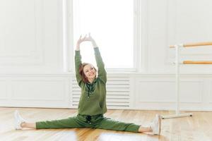 People, strength, fitness, gymnastics concept. Glad sportswoman with cheerful face, raises hands, spreads legs and does splits, stretches in studio. Young dancer has work out, dressed in tracksuit photo