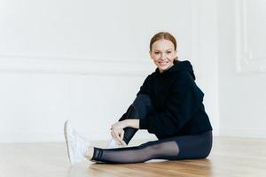 Indoor shot of pleased beautiful ginger woman has positive facial expression, dressed in black clothing, has rest on floor, satisfied after doing aerobic exercises, has sporty body, muscular legs photo