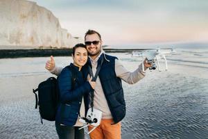Happy female and male travellers with bags and modern technologies for making photos and video of what they see. Couple in love spend vacations on seaside, try new equipment drone for first time