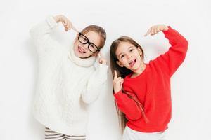 Joyful girls being in good mood, wear oversized sweater and spectacles, make cool sign, pose together against white background. Happy female children have fun. Childhood and happiness concept photo