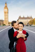 Affectionate male and female embrace each other, kiss, enjoy spending holidays in London, near Big Ben. Tourist couple go sightseeing outdoors in Great Britain. Tourism and relationship concept photo