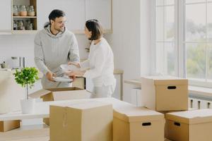 Photo of busy family couple unpack personal stuff from carton boxes, dressed in casual clothes, hold white plates, pose in spacious kitchen with modern furniture, surrounded with pile of packages