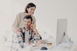 Happy young man holds mobile phone, discusses issues, manages home accounts with wife, pose in coworking space, sit in front of computer, discuss family expenses. Couple collaborate together photo