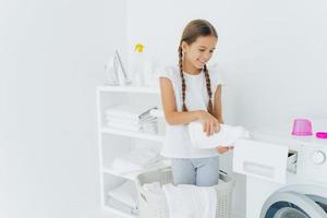 Small adorable busy girl stands in laundry basket, pours detergent in washing machine compartment, has glad expression, long hair combed in pigtails, does housework in laundry room. Cleanliness photo