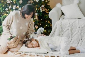 Attractive brunette smiling woman stands on knees with present boxes, little daughter sleeps near on pillow, has pleasant dreams, enjoy holiday. Winter time, New Year and Christmas. Rest concept photo